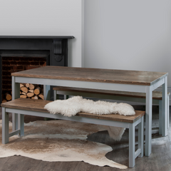 A grey and dark pine dining table with two matching benches set in a room with a fireplace, with a white furry throw on one of the benches.