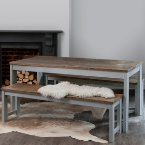 A grey and dark pine dining table with two matching benches set in a room with a fireplace, with a white furry throw on one of the benches.