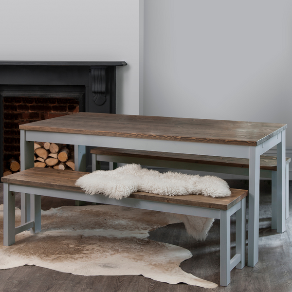 A grey and dark pine dining table with two matching benches set in a room with a fireplace, with a white furry throw on one of the benches.