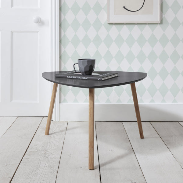 An extra large side table with a curved surface and three wooden legs, placed on a white floor against a wall with patterned wallpaper. The table is Storm Grey in color and has a book and a cup on top.
